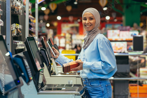 Self-service in a supermarket. A young Arab woman in a hijab is standing near the electronic cash registers and is paying for the goods in the store online through an application on her phone.