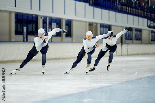 Three young athletes in sports uniform, skates and protective eyeglasses moving forwards along ice rink while taking part in competition