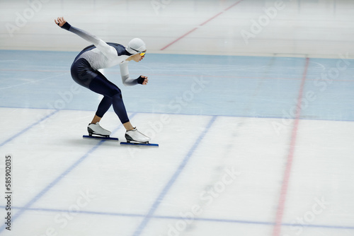 Young speed skater with legs bent in knees and right arm stretched behind back bending forwards while sliding down ice rink during race