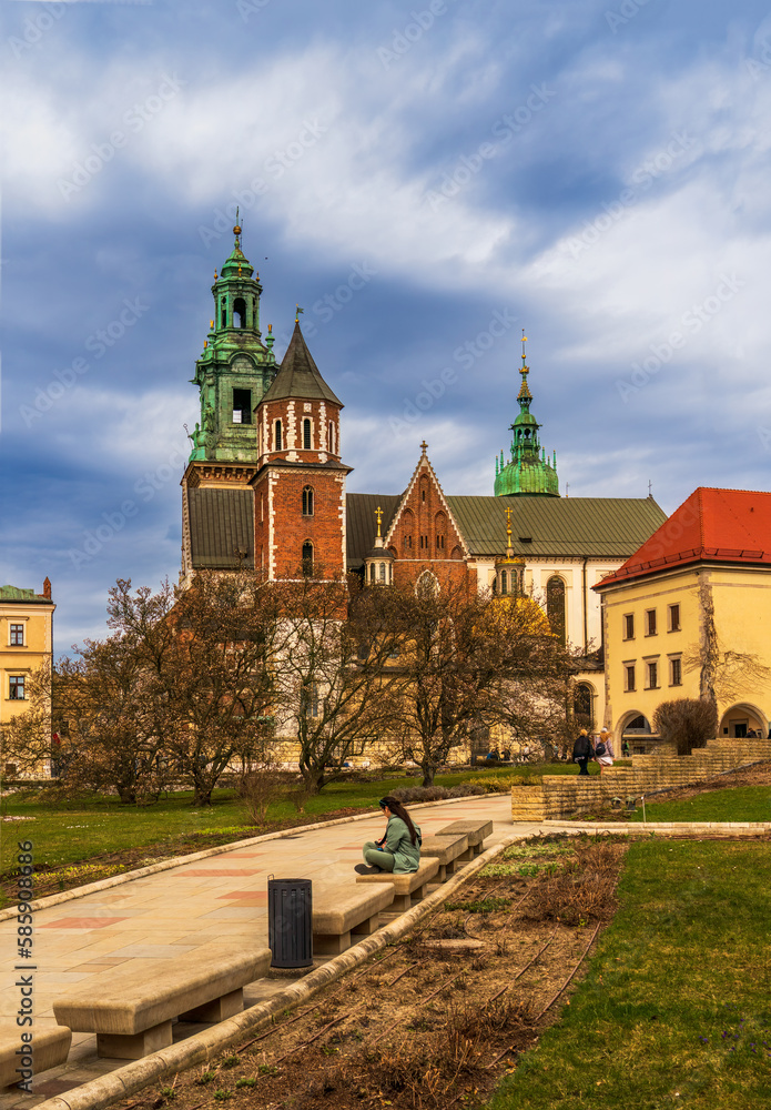 Historic Wawel Royal Castle in Krakow, Poland, on a spring day against a cloudy sky, magnolia trees with white flowers in the foreground, a tourist on a bench, traveling in European cities