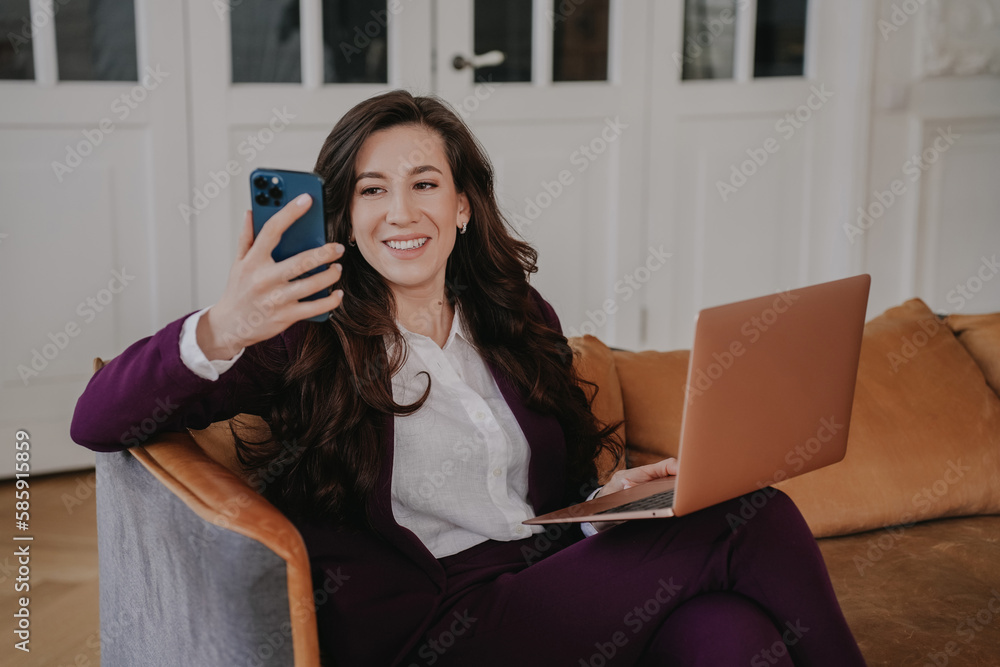 Brunette Spanish businesswoman sitting on chair with laptop holds phone toothy smiles makes video call at office. Successful hispanic entrepreneur remote works home. Young woman talks with friend. Brunette Spanish businesswoman sitting on chair with laptop holds phone toothy smiles makes video call at office. Successful hispanic entrepreneur remote works home. Young woman talks with friend.