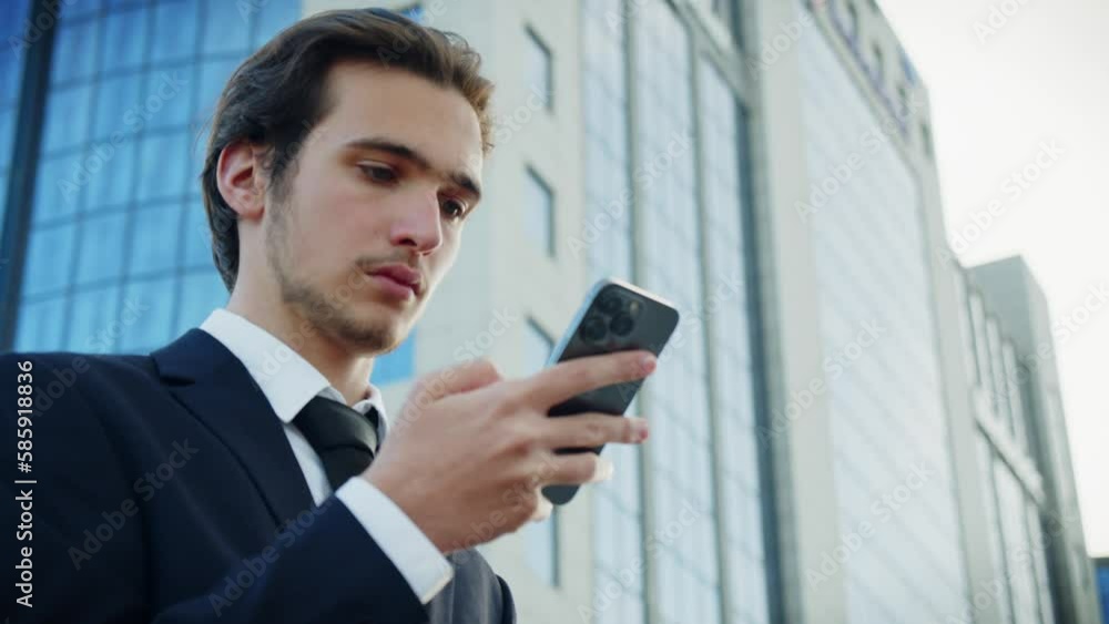 Confident businessman near a business center with a cell phone in hands. Successful young businessman in a blue suit is looking at a smartphone. Businessman is typing message on a mobile phone. Confident businessman near a business center with a cell phone in hands. Successful young businessman in a blue suit is looking at a smartphone. Businessman is typing message on a mobile phone.