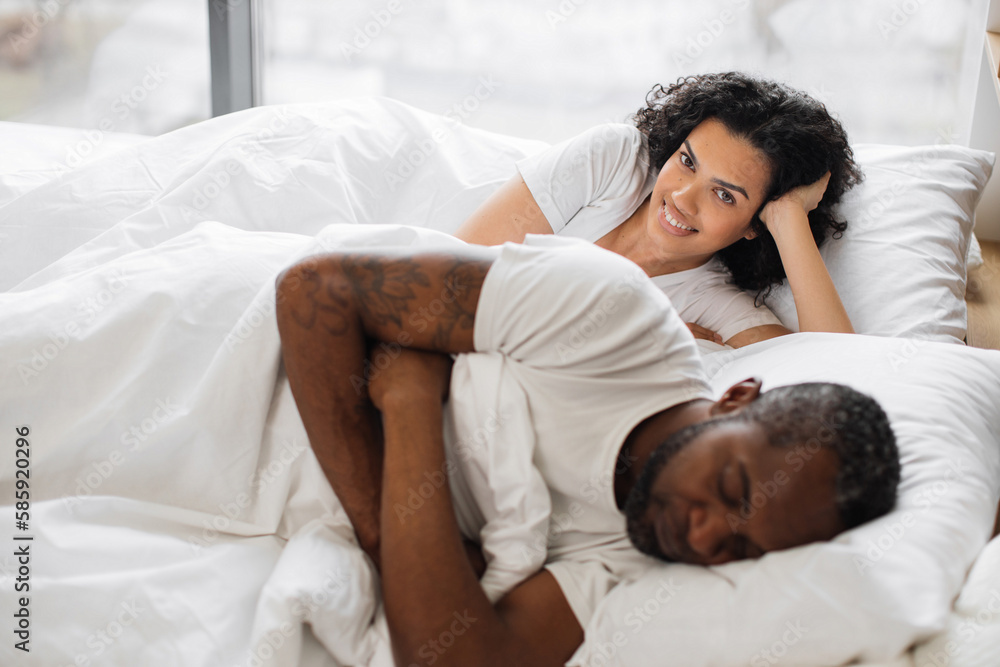 Pretty african american woman leaning on soft pillow already awake while her attractive husband still getting shuteye on bed in front of panoramic window. Happy couple spending morning together. Pretty african american woman leaning on soft pillow already awake while her attractive husband still getting shuteye on bed in front of panoramic window. Happy couple spending morning together.