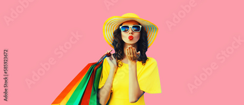 Portrait of beautiful happy young woman with shopping bags blowing her lips sending sweet air kiss wearing summer straw yellow hat on pink background