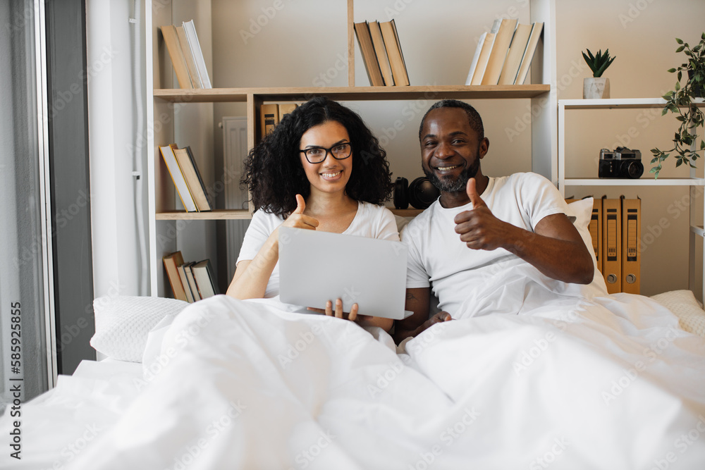 Cheerful multiracial couple holding portable computer and giving thumbs up sign while lying under white blanket at home. Romantic spouses encouraging to living comfortable tech lifestyles.