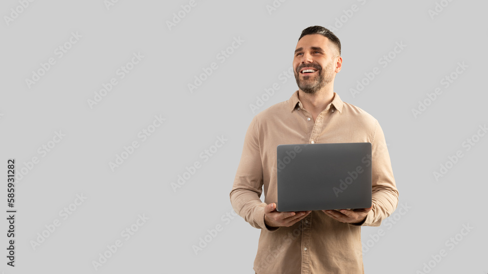 Positive confident middle aged businessman holding in hand opened laptop computer and looking at free space, panorama