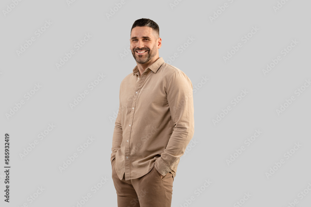 Fototapeta premium Portrait of positive middle aged man in shirt posing with hands in pockets smiling at camera, grey background