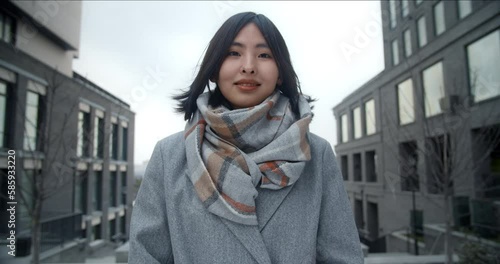 Portrait of a young beautiful Asian woman in a coat, standing between business buildings. Easy smile. Wind, snowfall.