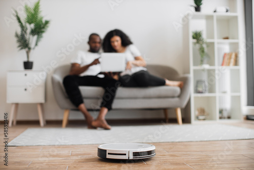 Automatic robot vacuum cleaning wooden floor while happy young couple resting on background. Two multicultural in love spending time for watching movie instead of households.