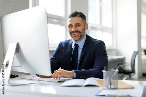Happy middle aged businessman in suit sitting in office and smiling at camera, working with modern computer, free space