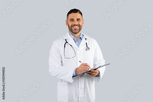 Happy middle aged man doctor in white coat with stethoscope, holding clipboard and smiling at camera, grey background