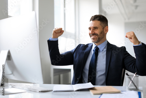 Business success. Excited middle aged businessman looking at computer and gesturing yes, shaking fists in joy in office
