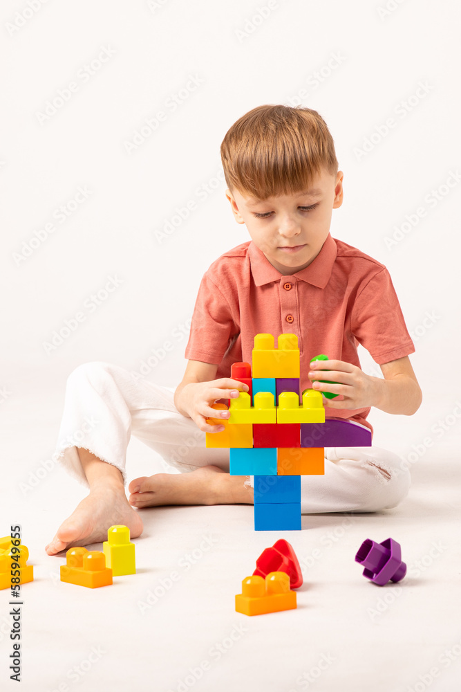 A happy child collects a tower of colored cubes. The preschooler sits down at the white background and assembles the construction set. Vertical photo. The boy smiles, pleased with the result. A happy child collects a tower of colored cubes. The preschooler sits down at the white background and assembles the construction set. Vertical photo. The boy smiles, pleased with the result.