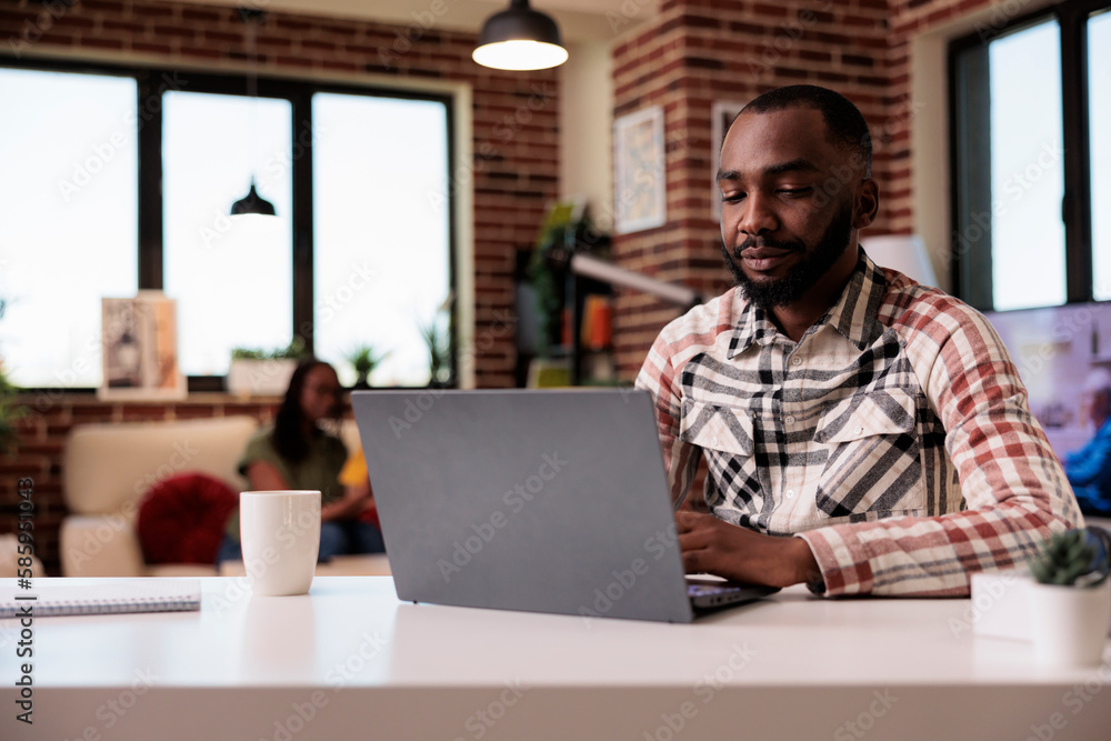 African american programmer writing code typing on laptop keyboard while girlfriend is reading a book in home living room. Male freelancer working remote focused on portable computer screen. African american programmer writing code typing on laptop keyboard while girlfriend is reading a book in home living room. Male freelancer working remote focused on portable computer screen.