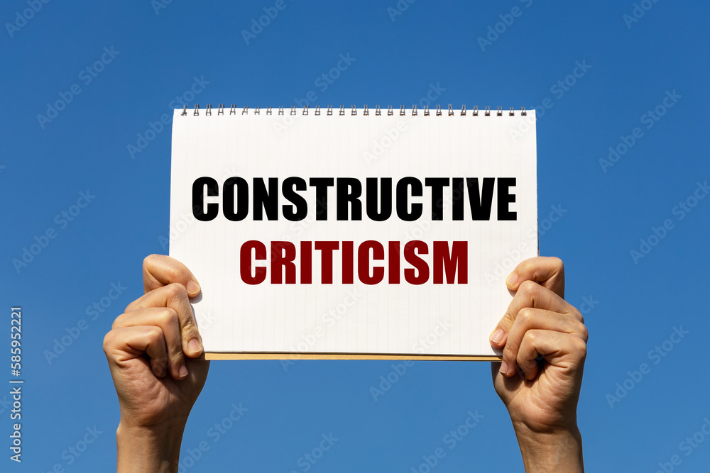 Constructive criticism text on notebook paper held by 2 hands with isolated blue sky background. This message can be used as business concept about constructive criticism. Constructive criticism text on notebook paper held by 2 hands with isolated blue sky background. This message can be used as business concept about constructive criticism.