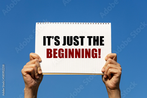 It's just the beginning text on notebook paper held by 2 hands with isolated blue sky background. This message can be used as business concept about it's just the beginning.