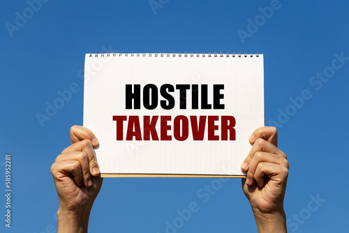 Hostile takeover text on notebook paper held by 2 hands with isolated blue sky background. This message can be used as business concept about hostile takeover. photo