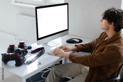 Male graphic designer or photographer working with empty screen computer at workspace, sitting at desk with equipment