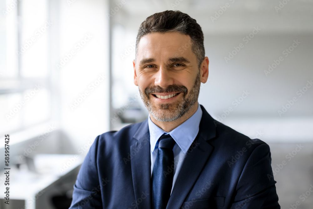Portrait of happy confident middle aged businessman in suit looking and smiling at camera, posing in office interior Portrait of happy confident middle aged businessman in suit looking and smiling at camera, posing in office interior