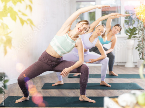 Positive women training standing yoga pose on black mat in light gym room with pot plants