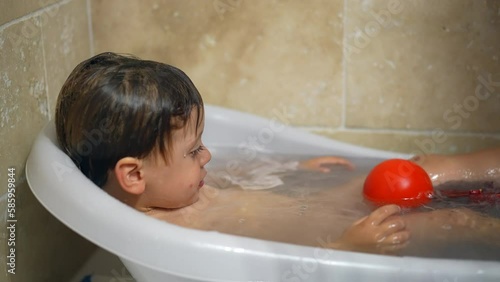 Cute baby bathing in small bathtub. Toddler boy washing routine bath tub