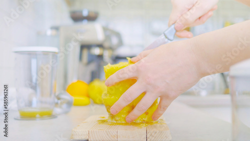 Young woman slicing a pineapple on a wooden cutting board in a kitchen surrounded by other fruits and a blender. She prepares a healthy, vitamin-rich and refreshing smoothie in one mixer.