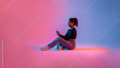 Amazing application. Happy black lady using smartphone, sitting on studio background in neon light, panorama, free space