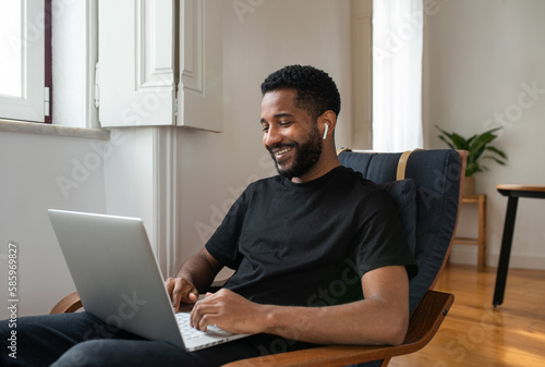 Young handsome cheerful black man working from home online remotely using his laptop. Looking at laptop screen, smiling.