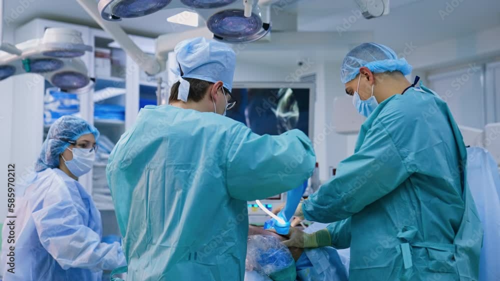 Rear view of a surgeon using device with mini camera. Assistant holding patient’s foot to help the doctor. Screen working at backdrop. Rear view of a surgeon using device with mini camera. Assistant holding patient’s foot to help the doctor. Screen working at backdrop.