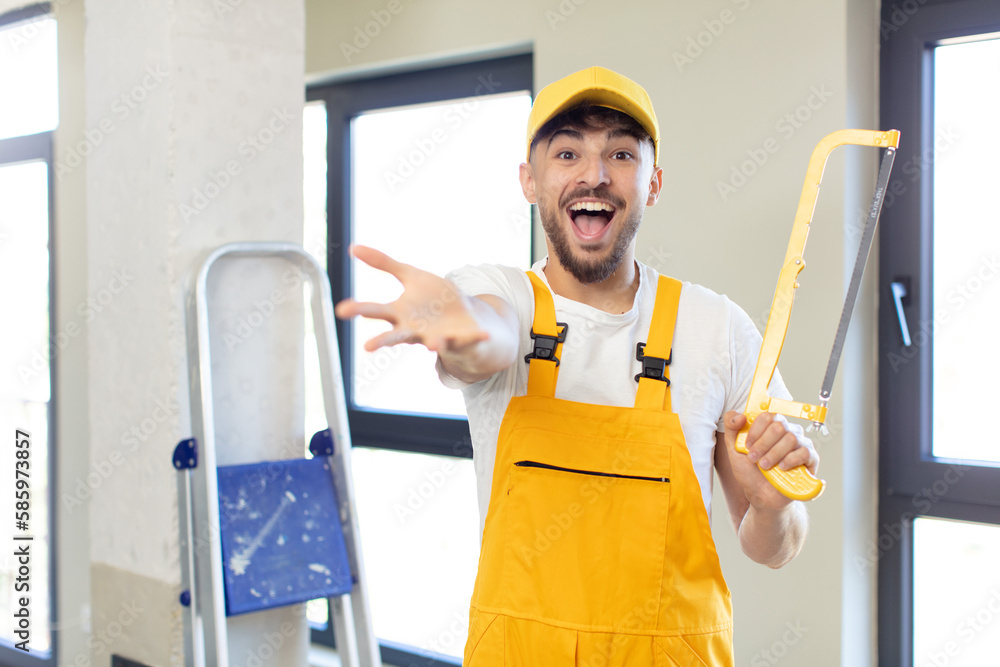 young handsome man smiling happily and offering or showing a concept. handyman with a saw young handsome man smiling happily and offering or showing a concept. handyman with a saw