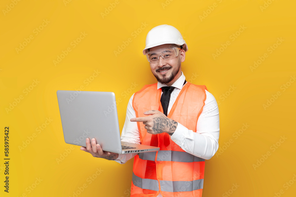 asian foreman in uniform advertises laptop and on yellow isolated background and points with his hand asian foreman in uniform advertises laptop and on yellow isolated background and points with his hand