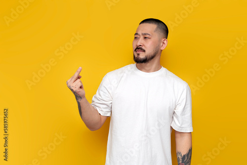 adult rude tattooed asian man in white t-shirt showing middle finger on yellow isolated background