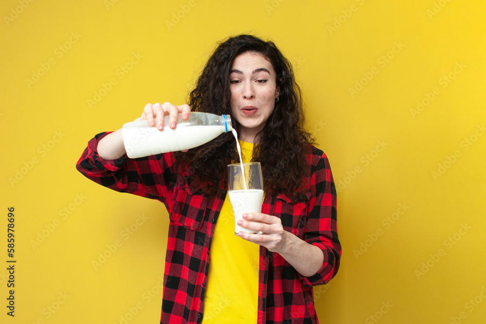 cheerful curly girl pours milk from a bottle into a glass on a yellow background and smiles cheerful curly girl pours milk from a bottle into a glass on a yellow background and smiles