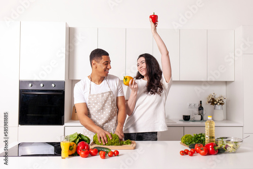 young happy multiracial couple preparing veggie vegetable and greens salad and dancing in white kitchen photo