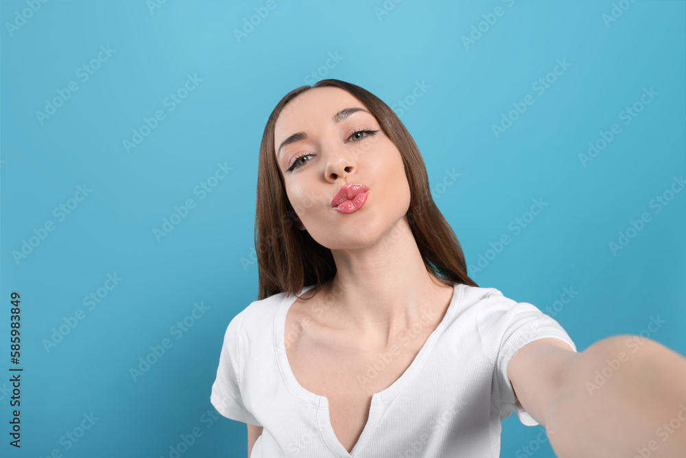 Beautiful young woman blowing kiss while taking selfie on light blue background Beautiful young woman blowing kiss while taking selfie on light blue background