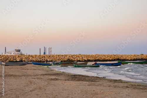 Colorful fishing boats with oars are docked on the beach next to a jetty made of large rocks © Dan Chis