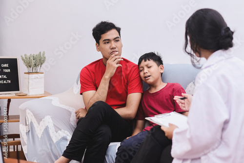 Professional female child psychologist working with crying school boy in a office. Woman makes notes and interview father about the condition of the boy on paper. Children's mental therapy. photo