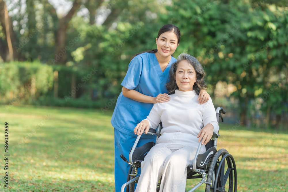 Young asian care helper with asia elderly woman on wheelchair relax together park outdoors to help and encourage and rest your mind with green nature. Looking camera. Young asian care helper with asia elderly woman on wheelchair relax together park outdoors to help and encourage and rest your mind with green nature. Looking camera.