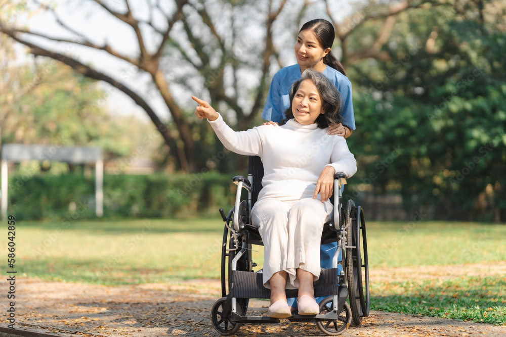 Young asian care helper with asia elderly woman on wheelchair relax together park outdoors to help and encourage and rest your mind with green nature. Pointing Young asian care helper with asia elderly woman on wheelchair relax together park outdoors to help and encourage and rest your mind with green nature. Pointing