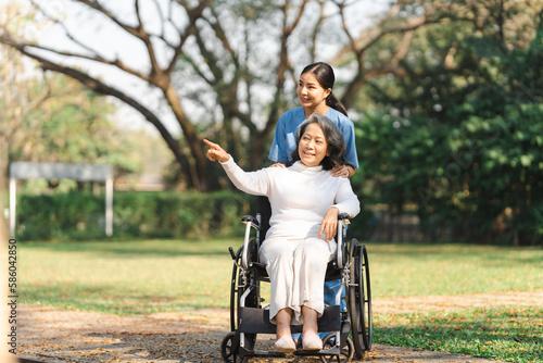 Young asian care helper with asia elderly woman on wheelchair relax together park outdoors to help and encourage and rest your mind with green nature. Pointing © NanSan