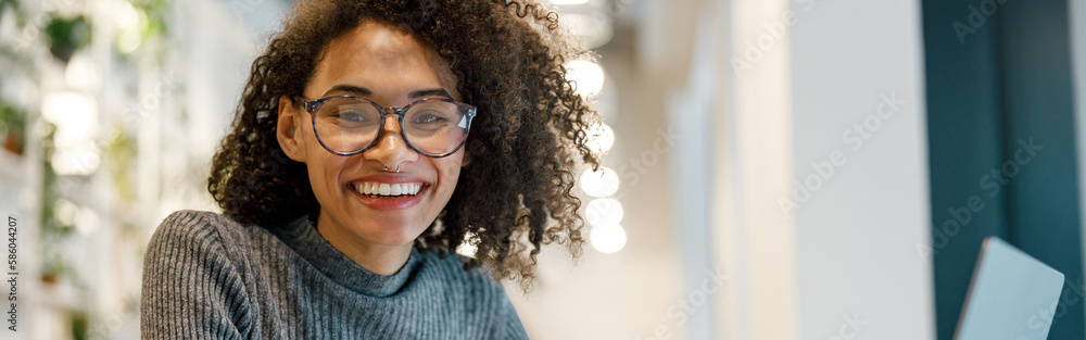 Smiling african woman freelancer working laptop while sitting in coworking and looking at camera - obrazy, fototapety, plakaty 
