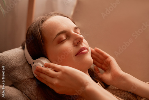 Side view portrait of relaxed young adult woman listening music while resting on couch at home, keeps eyes closed, touching her headphones, enjoying moment.