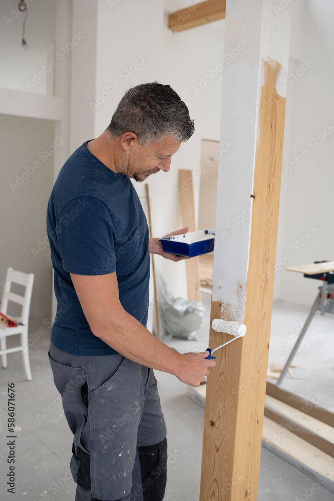 Middle-aged man with grey hair painting wooden beams in attic apartment, construction site with white paint Middle-aged man with grey hair painting wooden beams in attic apartment, construction site with white paint