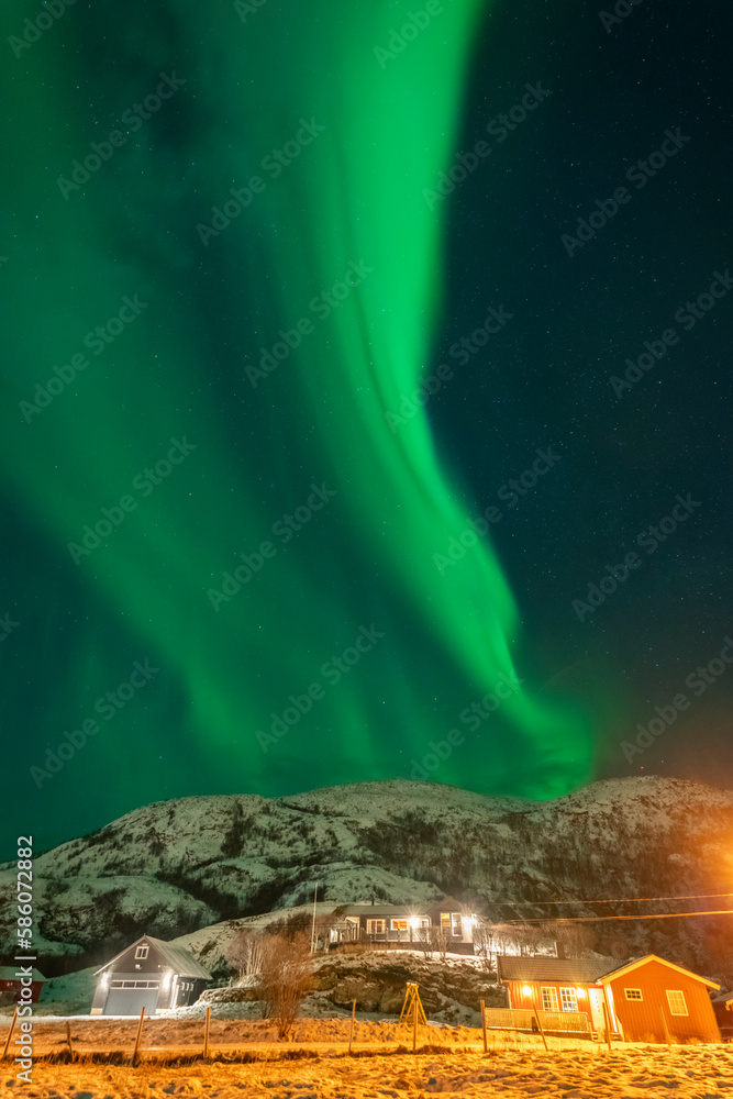 wunderbare Nordlichter über dem Dorf Hillesøy. Hell erleuchtete Häuser und Straßenlaternen bilden einen starken Kontrast zum dunklen Himmel mit der grünen Aurora Borealis. Winterstimmung in Norwegen