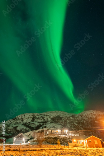 wunderbare Nordlichter über dem Dorf Hillesøy. Hell erleuchtete Häuser und Straßenlaternen bilden einen starken Kontrast zum dunklen Himmel mit der grünen Aurora Borealis. Winterstimmung in Norwegen