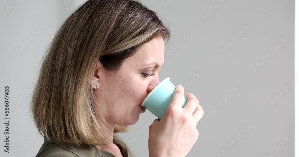 Serious woman drinks fresh water from paper cup in morning looking out window. Female employee quenches thirst standing on grey background slow motion Serious woman drinks fresh water from paper cup in morning looking out window. Female employee quenches thirst standing on grey background slow motion
