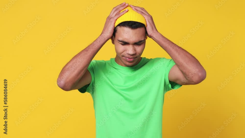 Displeased irritated sad angry young man of African American ethnicity wear green t-shirt hat closed eyes cover ears do not want to listen scream isolated on plain yellow background studio portrait Displeased irritated sad angry young man of African American ethnicity wear green t-shirt hat closed eyes cover ears do not want to listen scream isolated on plain yellow background studio portrait
