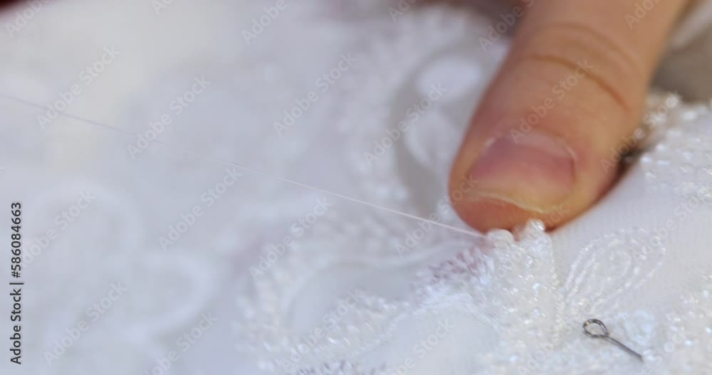 the female hand of a seamstress embroiders the details of a wedding dress with a needle and white thread at the factory. macro the female hand of a seamstress embroiders the details of a wedding dress with a needle and white thread at the factory. macro
