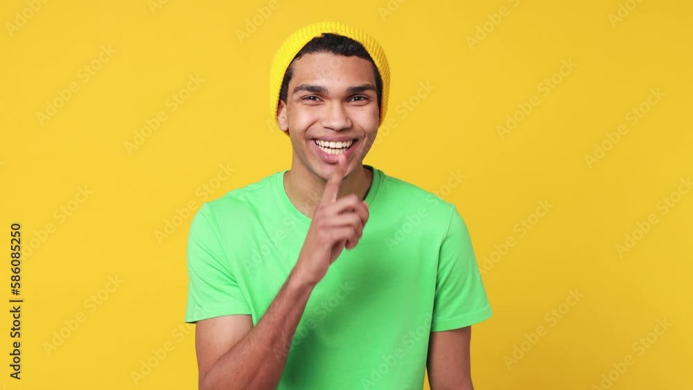 Secret young man of African American ethnicity wearing green t-shirt hat look aside say do not talk hush be quiet with finger on lips shhh gesture isolated on plain yellow background studio portrait Secret young man of African American ethnicity wearing green t-shirt hat look aside say do not talk hush be quiet with finger on lips shhh gesture isolated on plain yellow background studio portrait