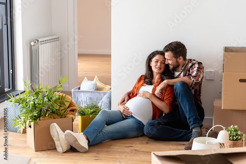 Beautiful young couple expecting a baby just moved into an empty apartment, sitting among cardboard boxes making plans for the future. New beginnings
